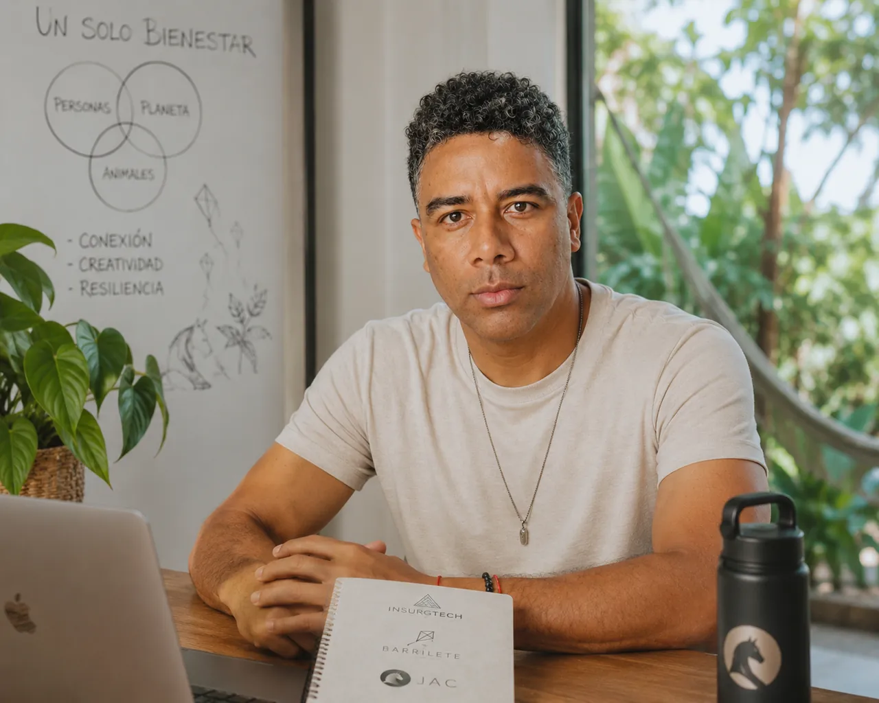 Eduardo Rengifo sentado a una mesa de trabajo, con una libreta que lleva los logos de Insurgtech, Barrilete y JAC; detrás, un tablero con el dibujo de Un Solo Bienestar